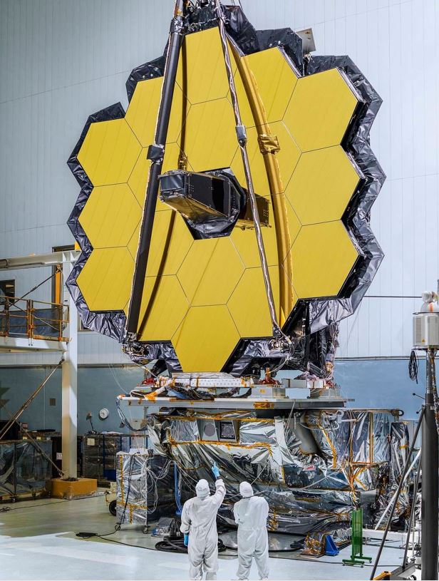The James Webb Space Telescope (JWST) team in the clean room standing underneath the
                    massive primary mirror.